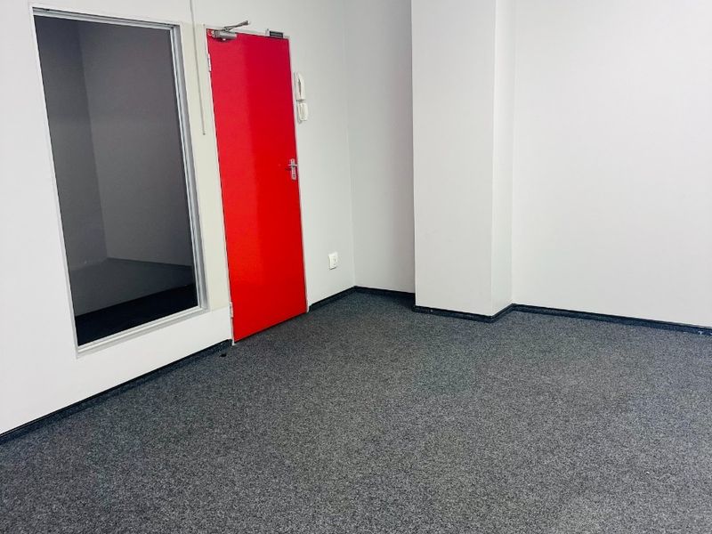 A clean, vacant office space featuring grey carpet flooring, a suspended ceiling, and a distinctive red door with an adjacent glass partition.