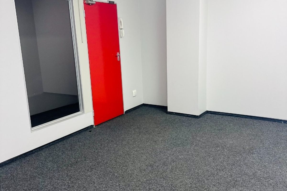 A clean, vacant office space featuring grey carpet flooring, a suspended ceiling, and a distinctive red door with an adjacent glass partition.