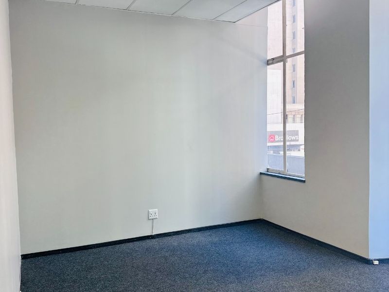 A clean, empty office space featuring carpeted flooring, a suspended ceiling with recessed lighting, and a large window providing natural light.