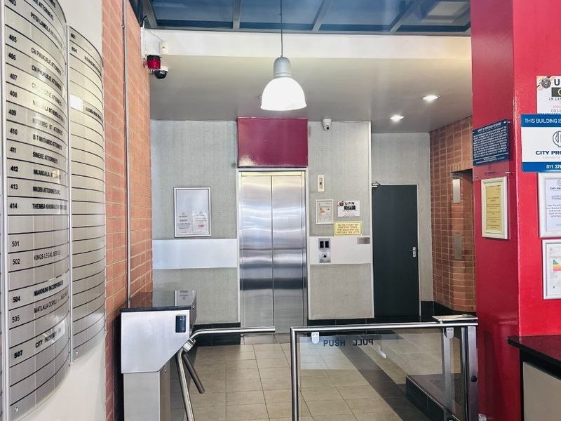 A professional office building lobby featuring a secure turnstile entrance, directory board, and elevator access with modern tiled flooring and suspended ceiling.