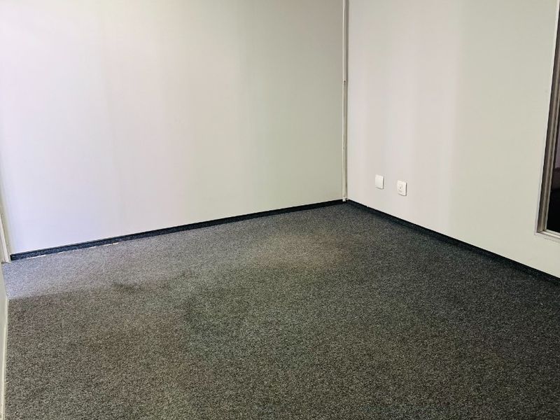 A vacant office space featuring neutral walls, a suspended ceiling, and grey carpet flooring with visible signs of wear.