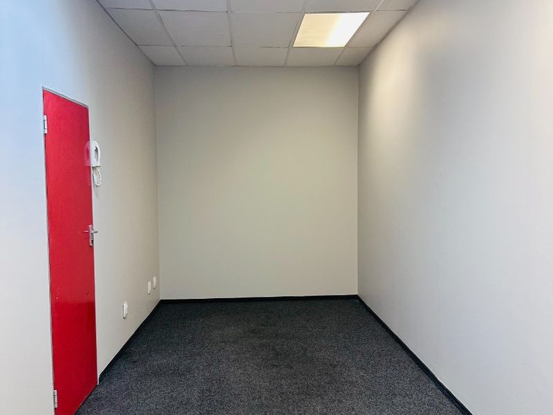 A clean, empty office space featuring neutral walls, dark carpet flooring, and a suspended ceiling with recessed lighting.