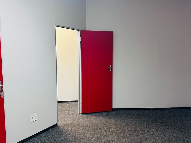 A clean, empty office space featuring grey carpet flooring, a suspended ceiling, and a distinctive red internal door.
