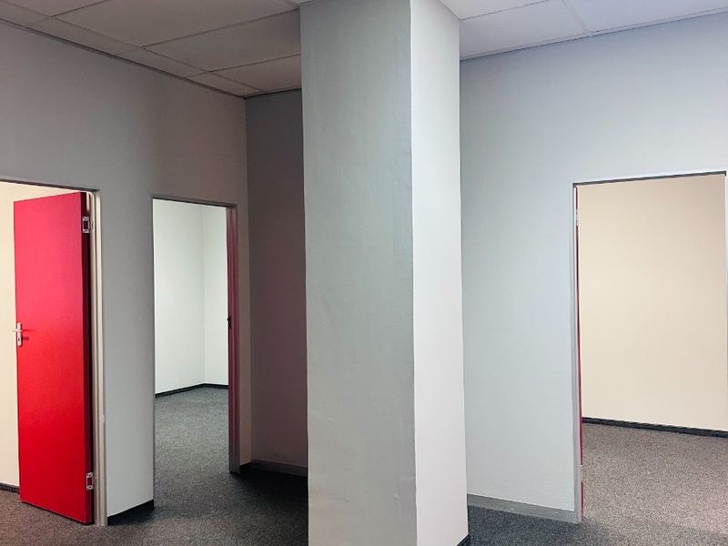 An empty office space featuring a central structural column, suspended ceiling with recessed lighting, and grey carpet flooring with multiple doorways.