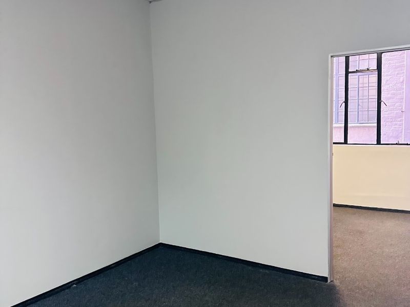 A vacant office space featuring neutral walls, carpet flooring, and a suspended ceiling, with an adjacent room visible through the doorway.