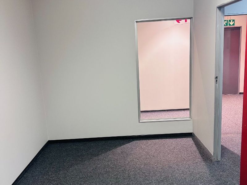 An empty office space featuring grey carpet flooring, a suspended ceiling, and an internal glass partition window looking into the adjacent corridor.