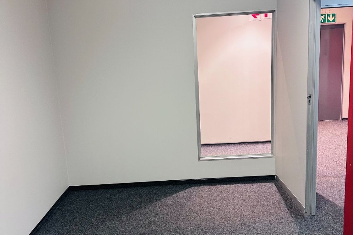 An empty office space featuring grey carpet flooring, a suspended ceiling, and an internal glass partition window looking into the adjacent corridor.