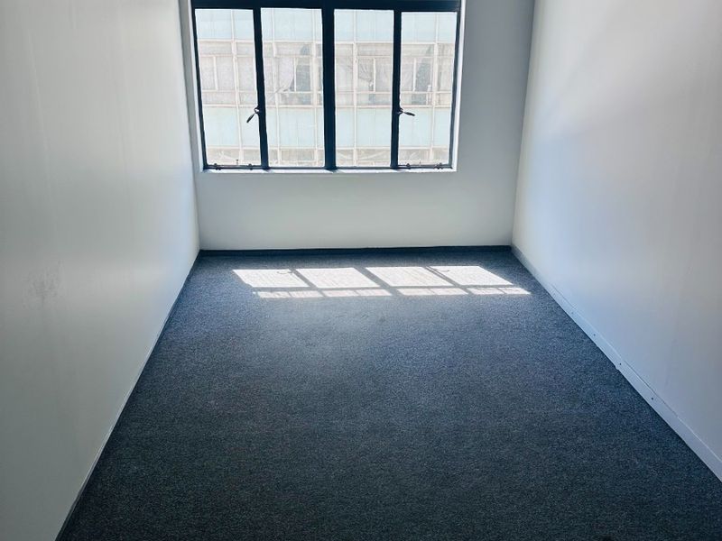 A bright, empty office space featuring neutral walls, carpeted flooring, and a large multi-pane window providing natural light.