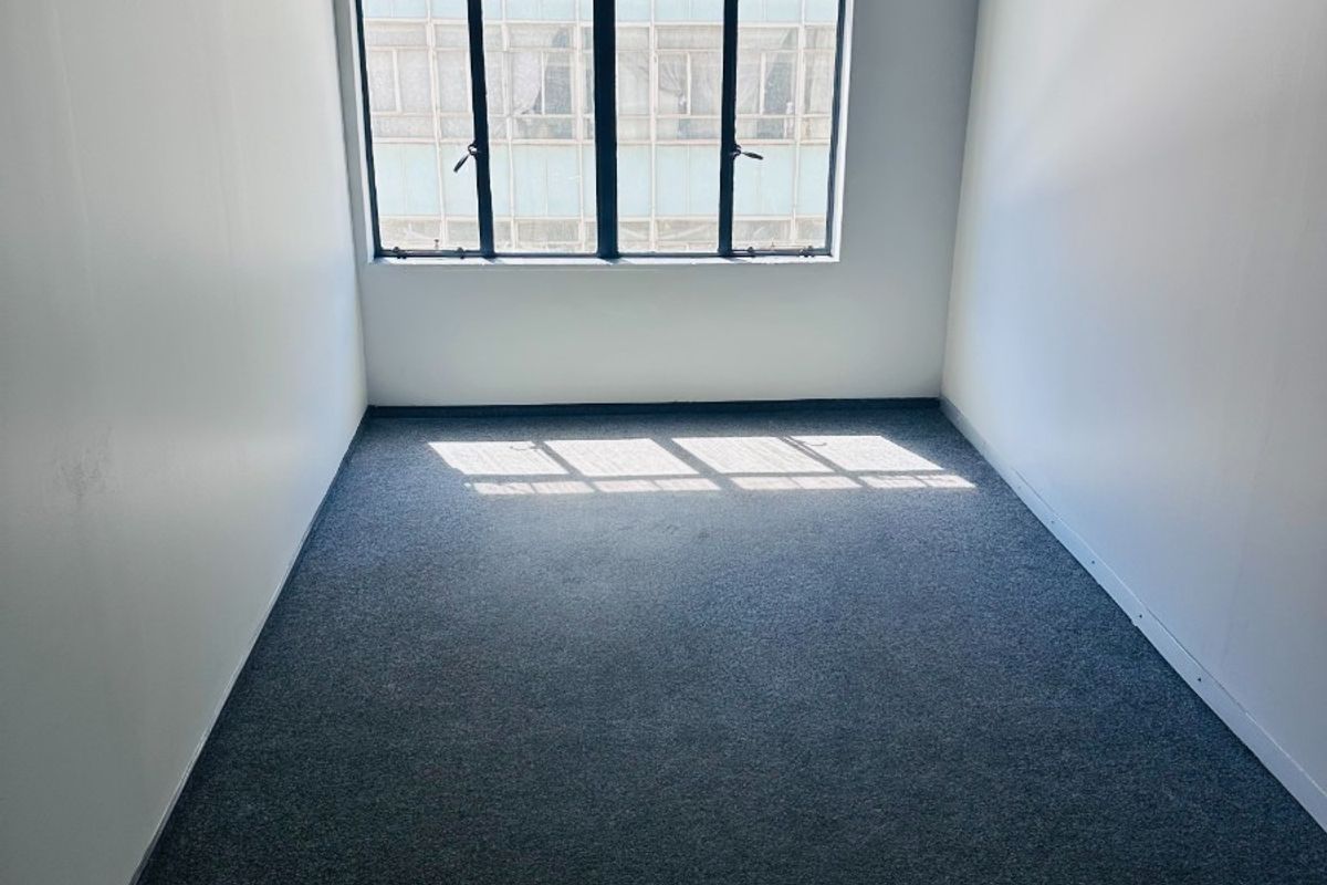 A bright, empty office space featuring neutral walls, carpeted flooring, and a large multi-pane window providing natural light.