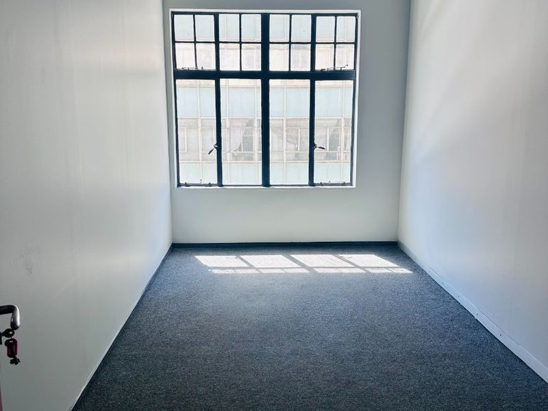 A bright, empty office space featuring neutral walls, carpeted flooring, and a large window providing natural light.