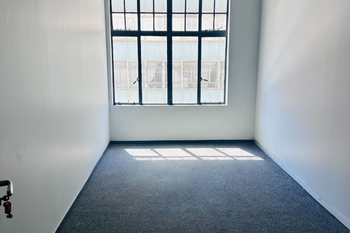 A bright, empty office space featuring neutral walls, carpeted flooring, and a large window providing natural light.