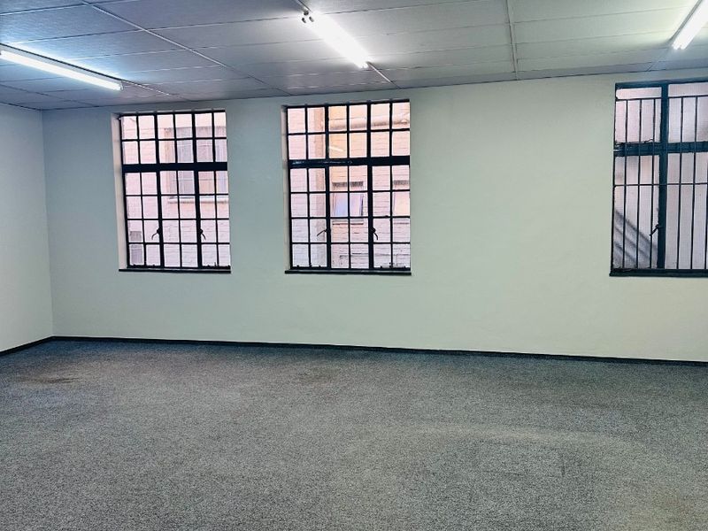 An empty, open-plan office space featuring a suspended ceiling, fluorescent lighting, and large steel-framed windows with security bars.