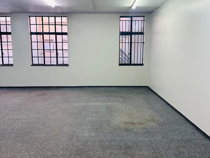 An empty office space featuring a suspended ceiling, fluorescent lighting, and security bars on the windows, with visible wear on the carpet flooring.