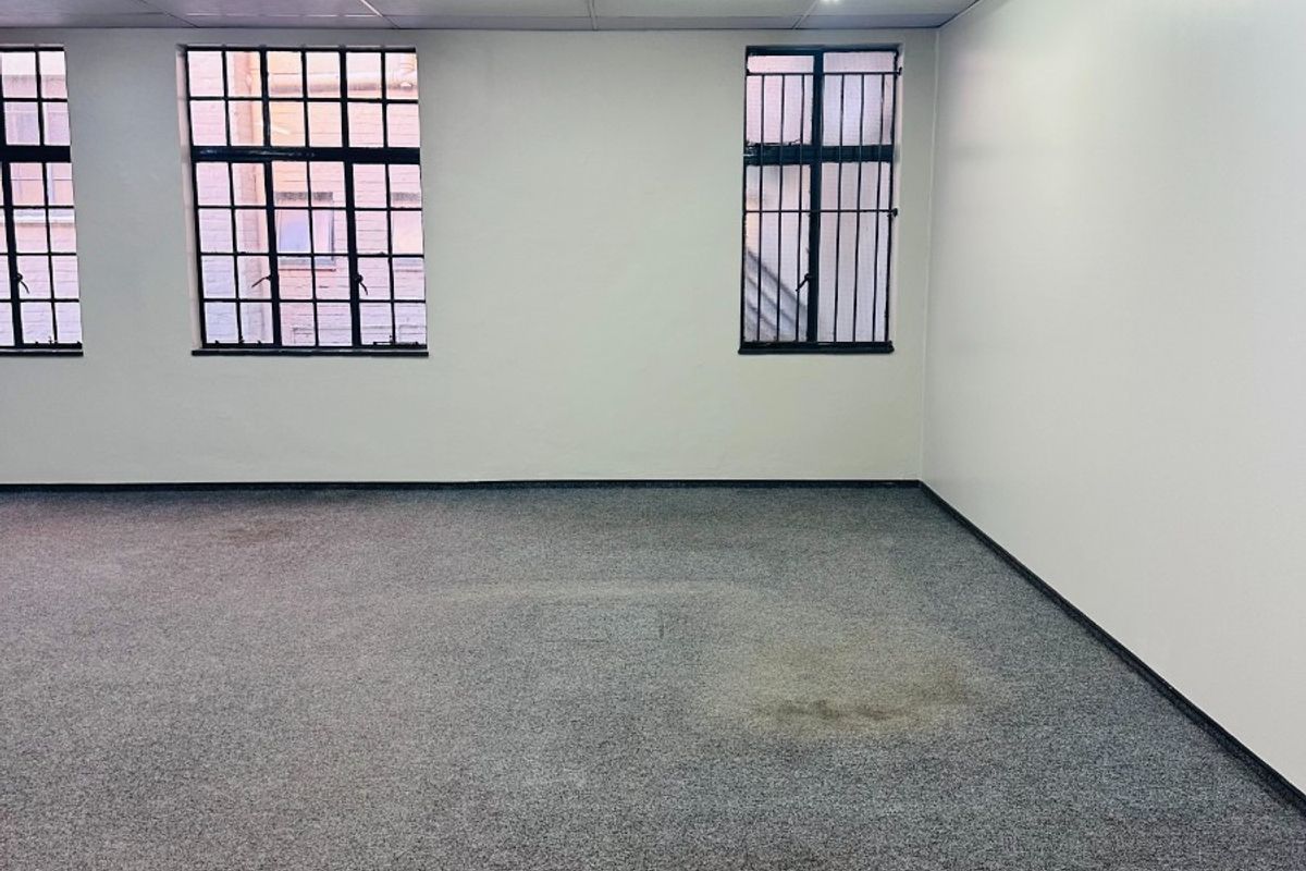 An empty office space featuring a suspended ceiling, fluorescent lighting, and security bars on the windows, with visible wear on the carpet flooring.