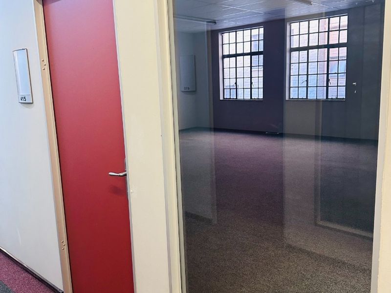 An empty office space featuring a glass partition wall, carpeted flooring, and large multi-pane windows providing natural light.