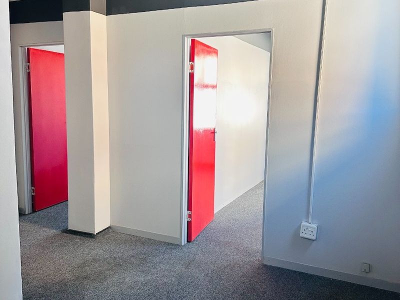 An empty office space featuring grey carpet flooring, exposed electrical conduit, and distinctive red interior doors leading to adjacent rooms.