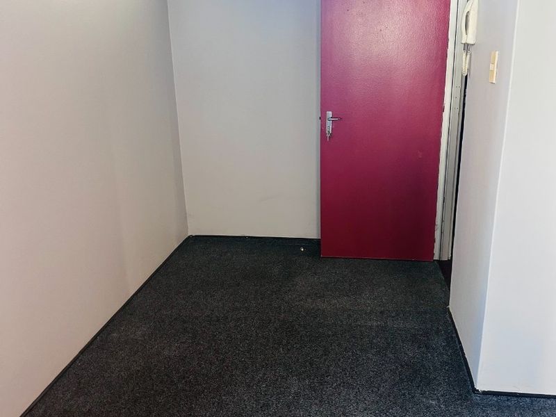 A compact, empty office space featuring grey carpet flooring, a high-level window for natural light, and a red interior door.