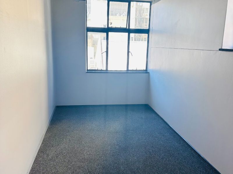A vacant office space featuring grey carpeting, neutral walls with a blue accent border, and a large window providing natural light.
