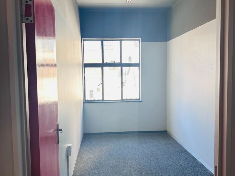 A compact, empty office space featuring grey carpet flooring, a multi-pane window for natural light, and a simple overhead fluorescent light fixture.