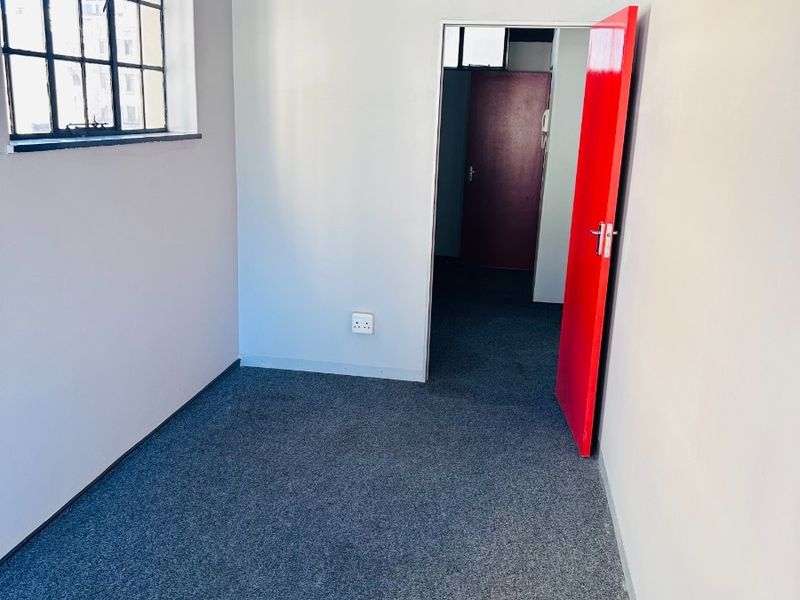 A vacant office space featuring industrial-style steel windows, grey carpeting, and a distinctive red door, offering a clean canvas for professional use.