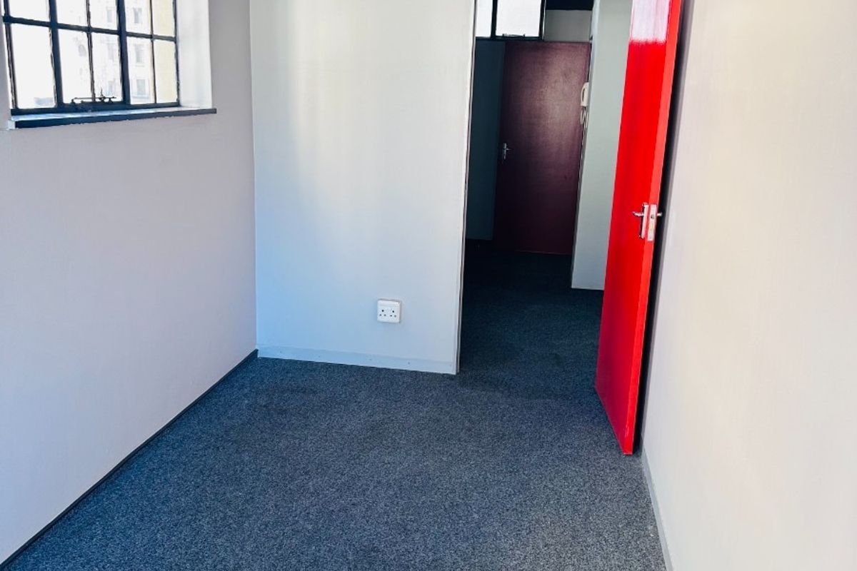 A vacant office space featuring industrial-style steel windows, grey carpeting, and a distinctive red door, offering a clean canvas for professional use.