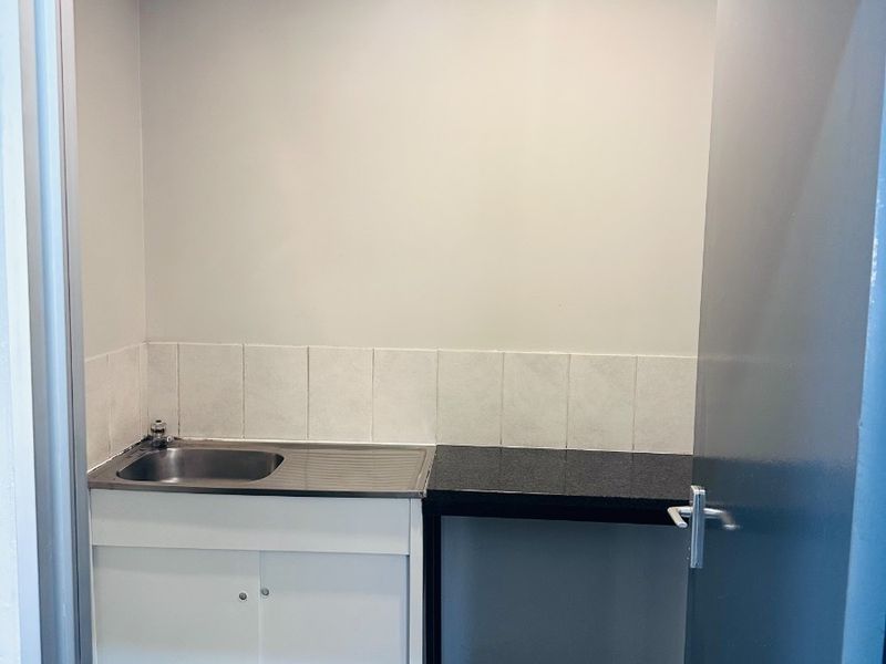 A compact office kitchenette featuring a stainless steel sink, tiled splashback, and dark laminate countertop with built-in base cabinetry.