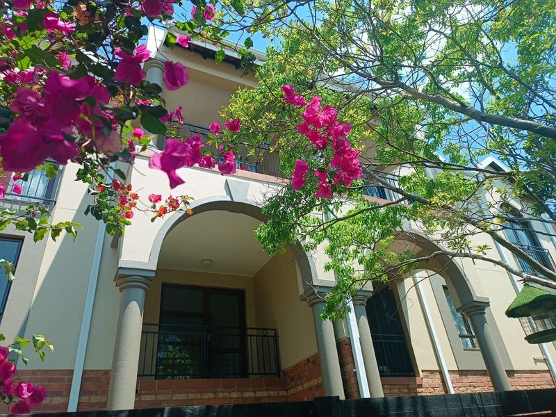 A view of the apartment building facade featuring arched balconies, decorative columns, and a brick-accented base, framed by lush flowering bougainvillea.