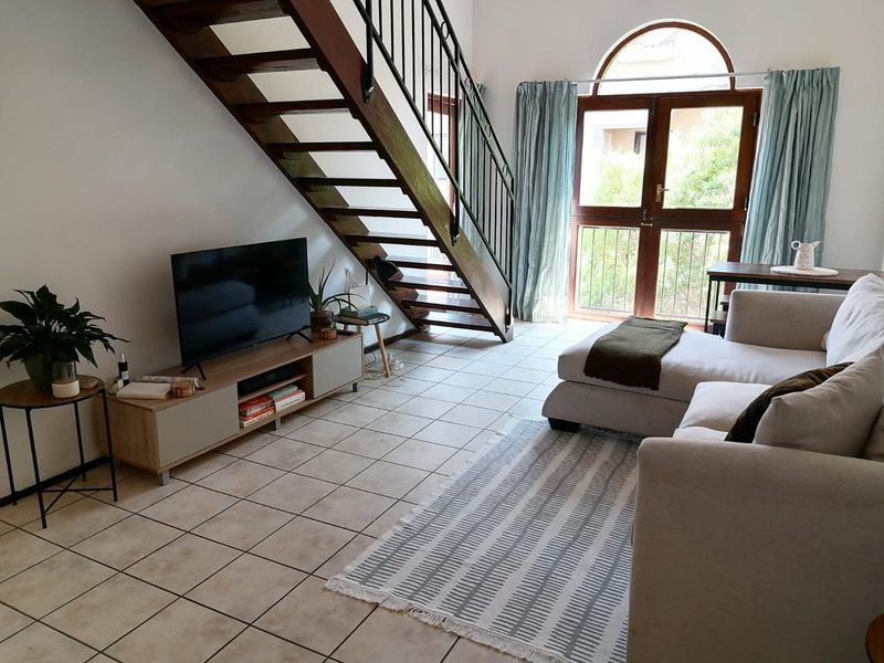 A bright living room featuring an open-tread timber staircase with wrought iron balustrade, ceramic tiled flooring, and an arched glass door leading outside.