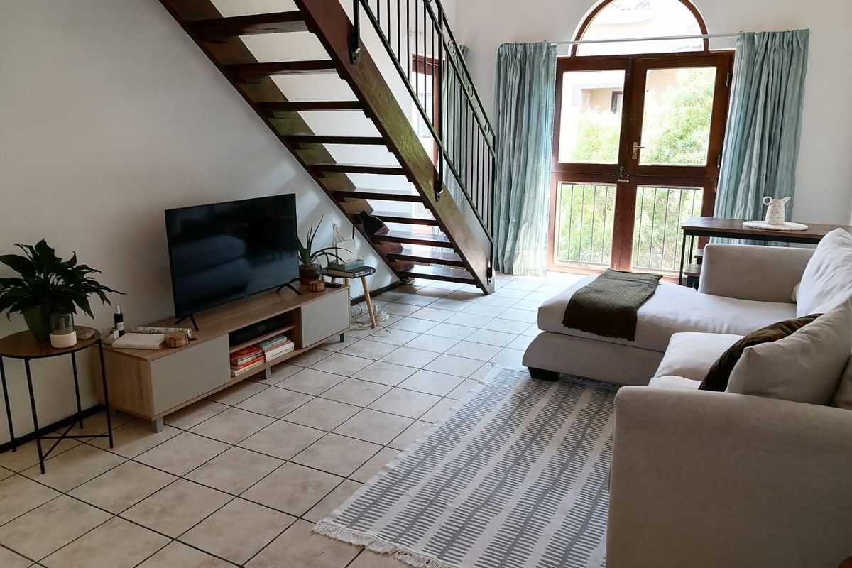 A bright living room featuring an open-tread timber staircase with wrought iron balustrade, ceramic tiled flooring, and an arched glass door leading outside.