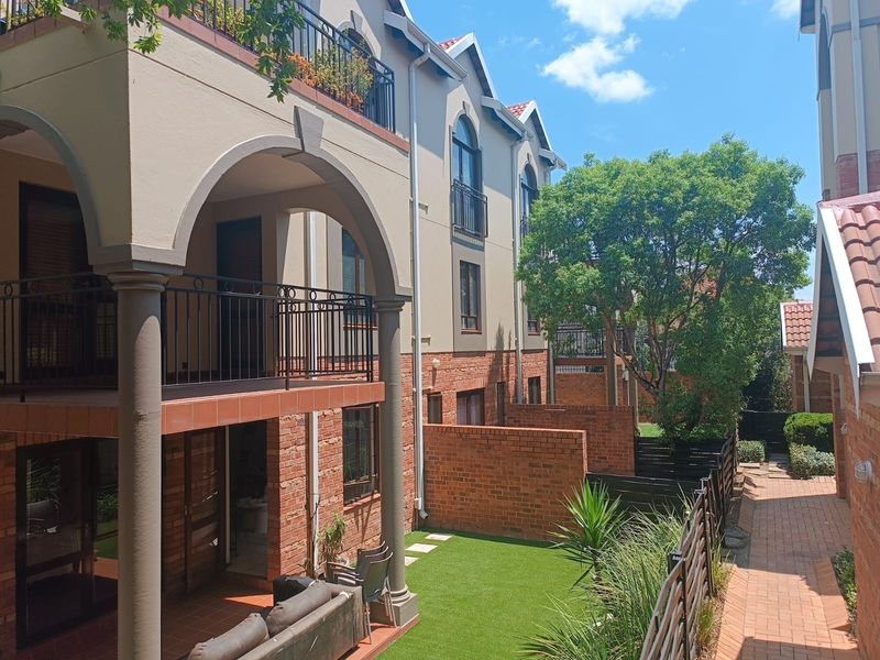 A view of the rear exterior of the apartment complex, featuring arched covered patios, brick finishes, and a well-maintained garden area with paved walkways.
