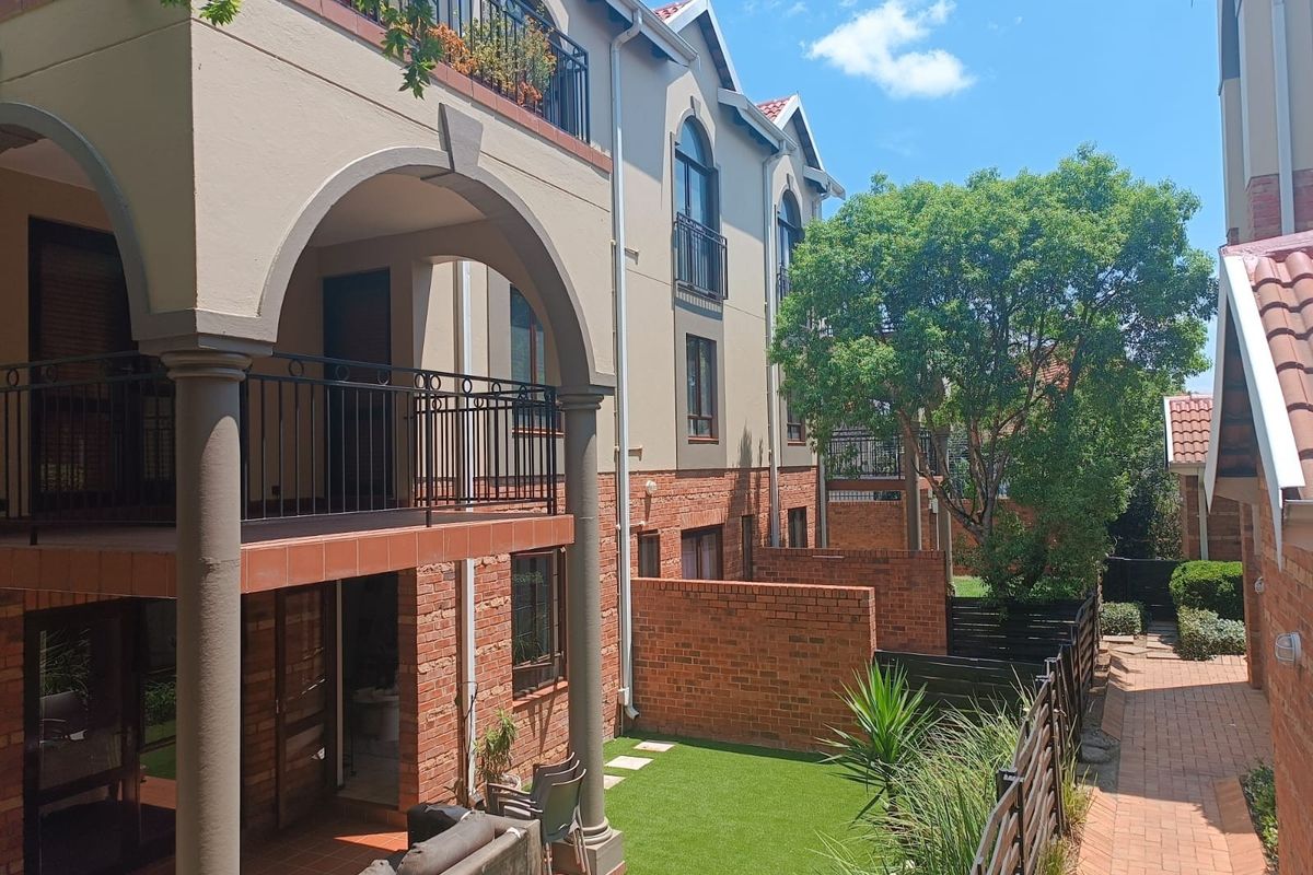 A view of the rear exterior of the apartment complex, featuring arched covered patios, brick finishes, and a well-maintained garden area with paved walkways.
