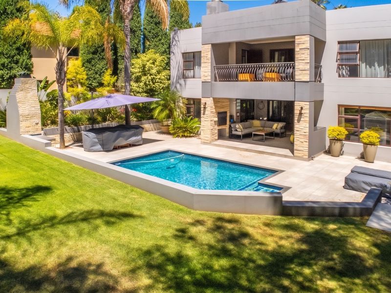 A well-maintained rear exterior featuring a modern two-story home, a sparkling swimming pool, a covered patio with built-in braai, and a landscaped garden.