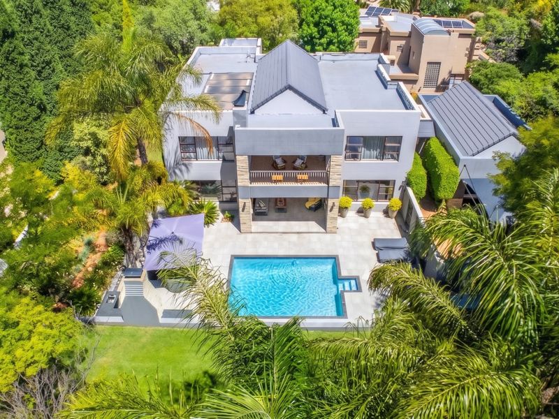 Aerial view of a modern multi-story residence featuring a swimming pool, spacious tiled patio, upper-level balcony, and lush landscaped garden surroundings.