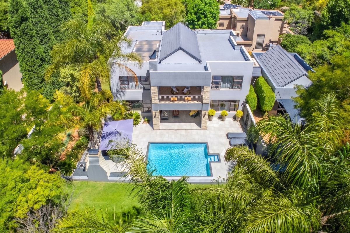Aerial view of a modern multi-story residence featuring a swimming pool, spacious tiled patio, upper-level balcony, and lush landscaped garden surroundings.