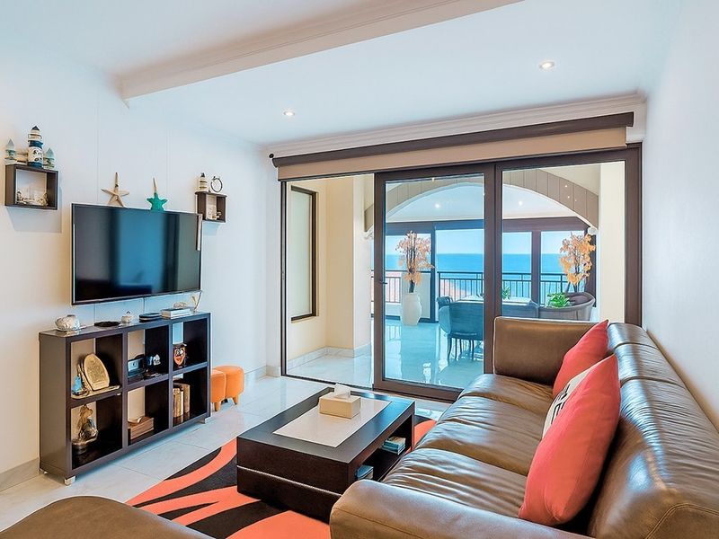 A bright living room featuring tiled floors, recessed lighting, and large sliding glass doors that open onto a balcony with ocean views.
