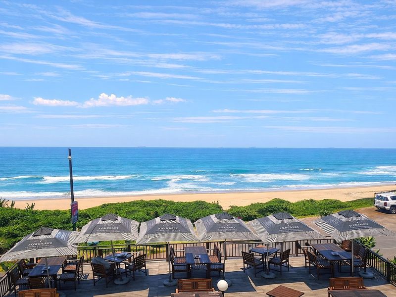 A scenic elevated view overlooking the ocean, sandy beach, and coastal vegetation, featuring a spacious outdoor timber deck area.