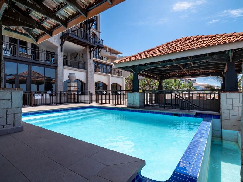 A well-maintained swimming pool area featuring a covered gazebo, stone-clad pillars, and modern metal safety railings within an apartment complex setting.