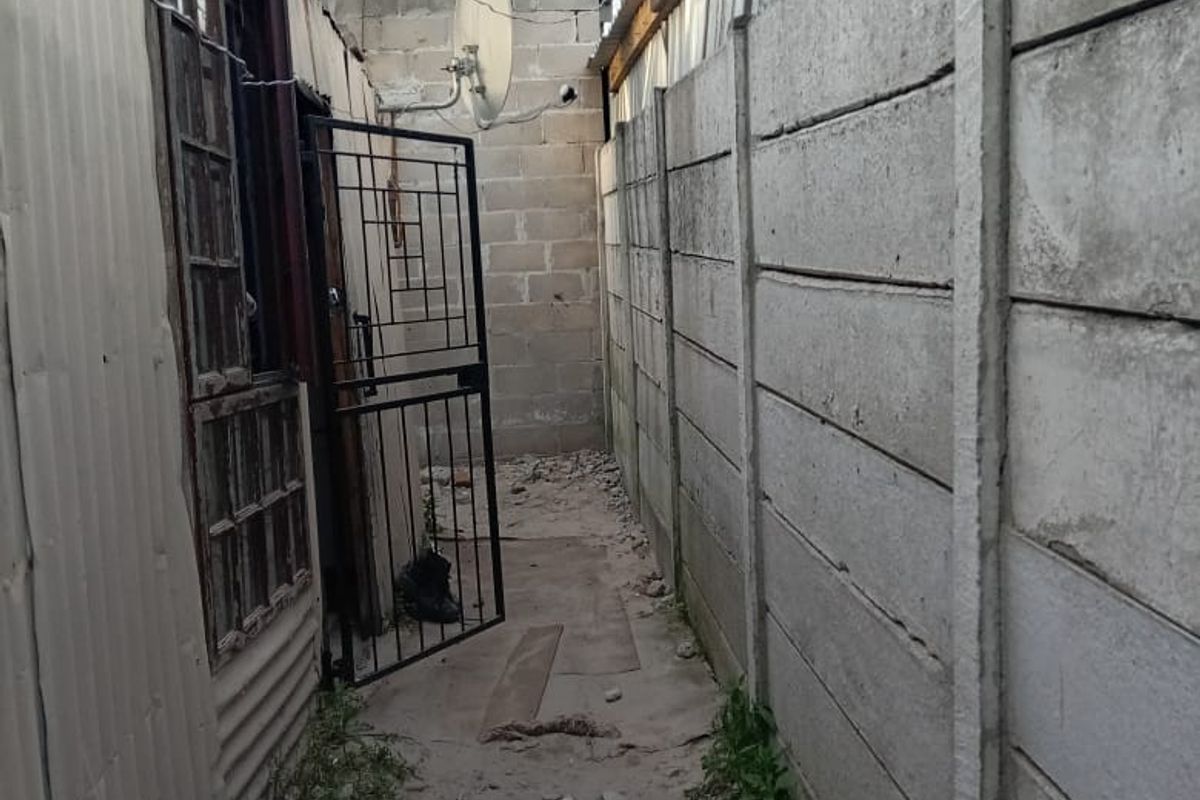 A narrow side passage between a corrugated metal structure and a precast concrete boundary wall, featuring a security gate and unpaved ground.