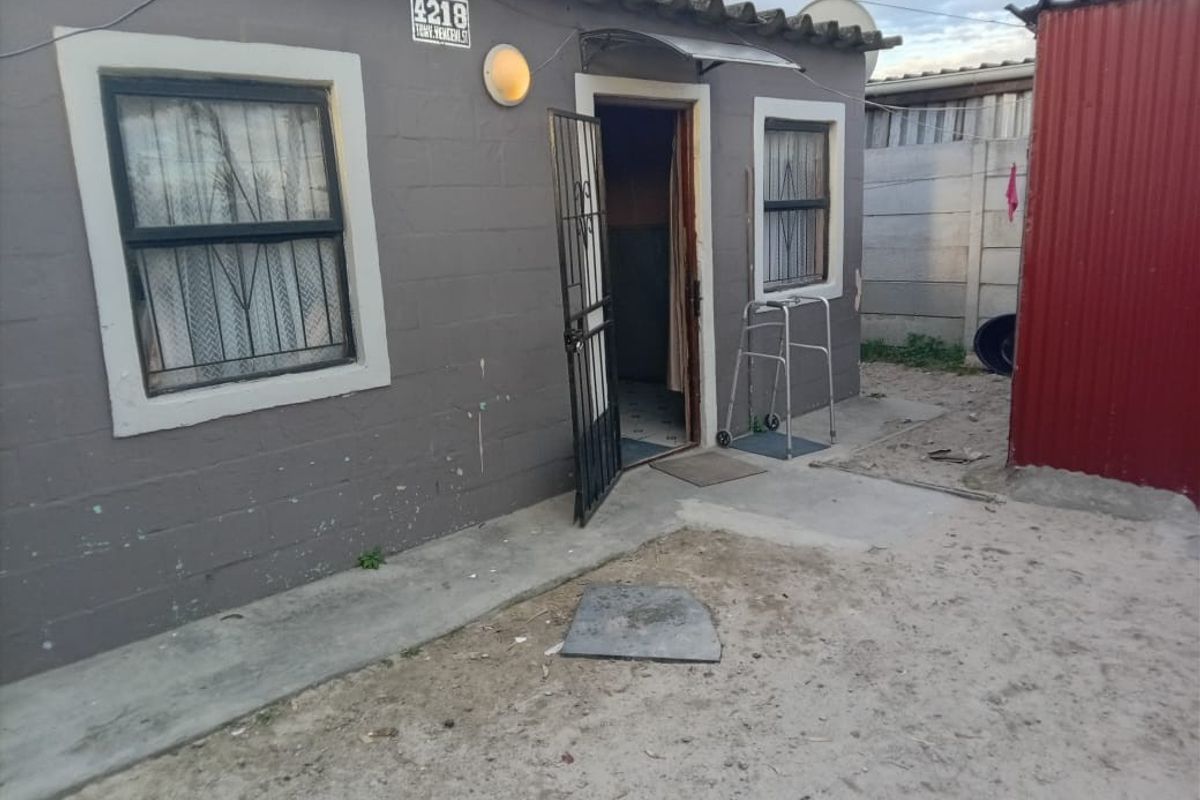 Front exterior view of a single-story concrete block house featuring a security gate, exterior lighting, and a basic concrete apron entrance.