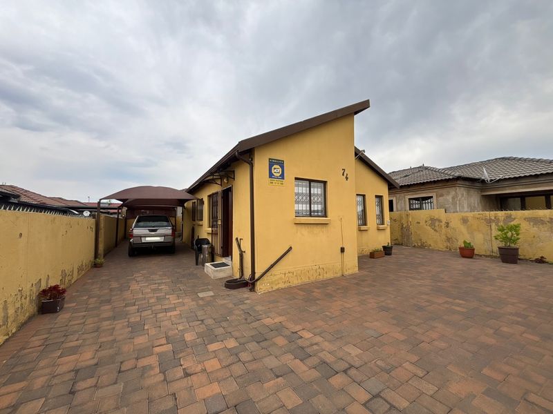 Front exterior view of a single-story house featuring a paved driveway, carport, and perimeter walling with visible exterior paint wear.