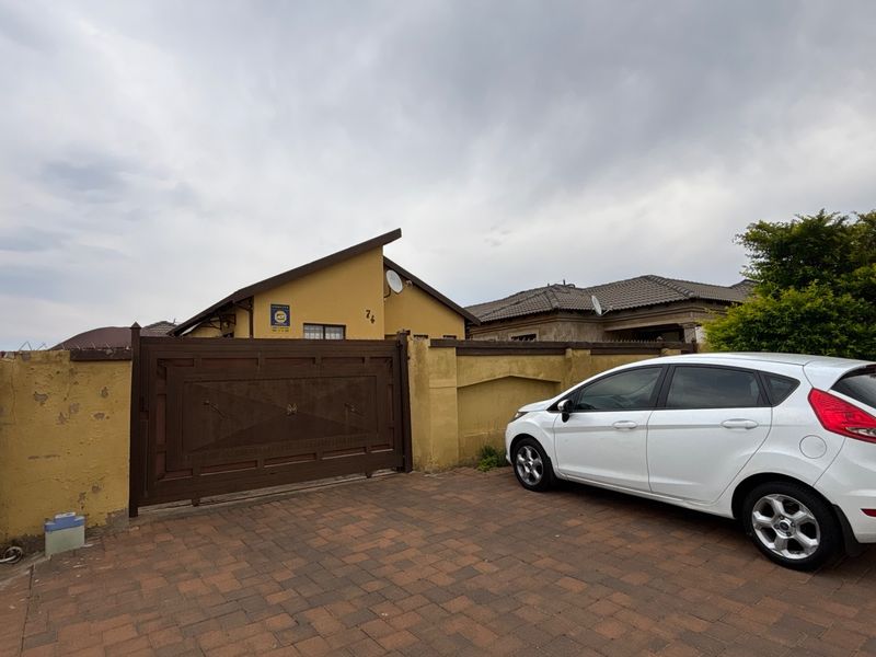 Front exterior view of the property featuring a paved driveway, a secure metal gate, and a perimeter wall with visible exterior finish wear.