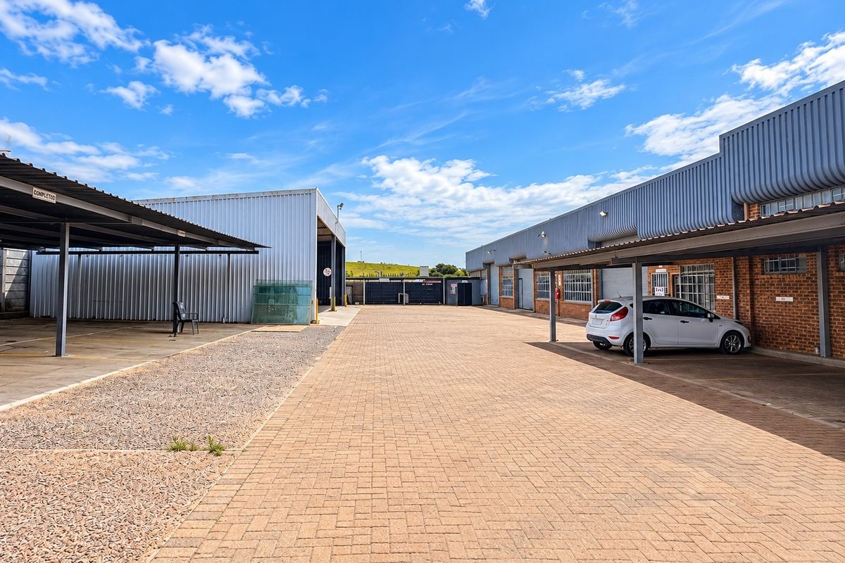 A spacious commercial exterior area featuring paved driveway, covered parking bays, and a combination of brick and corrugated metal building facades.