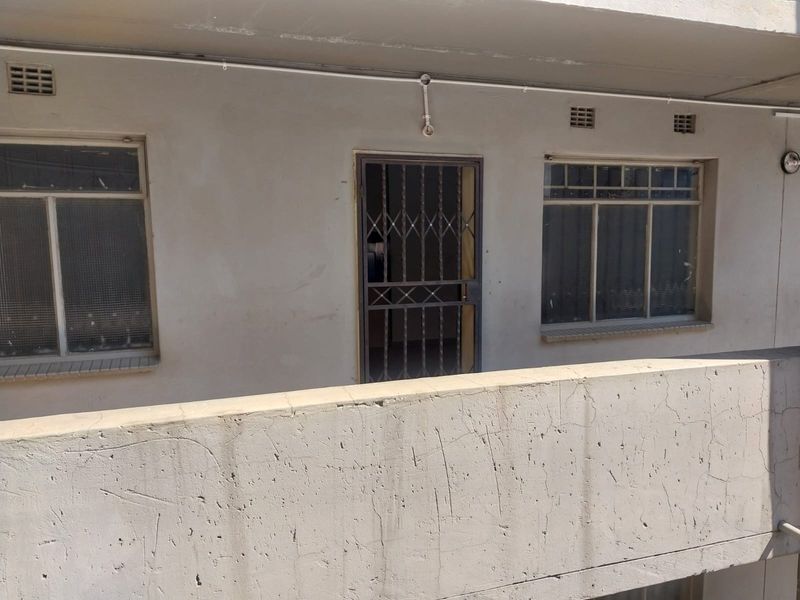 Exterior view of an apartment balcony featuring a concrete balustrade, security gate, and steel-framed windows with visible signs of weathering.