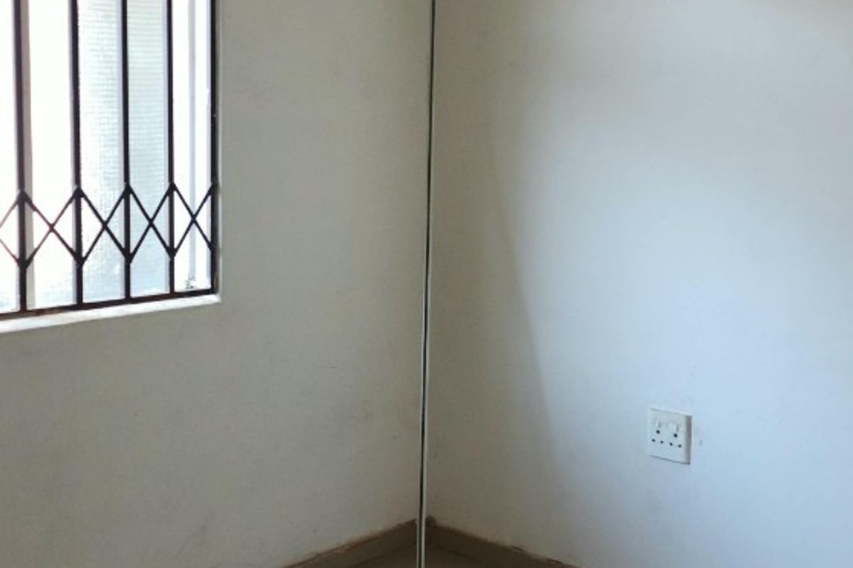 A vacant bedroom featuring tiled flooring, a security-gated window, and basic electrical fixtures, ready for a fresh coat of paint and new finishes.