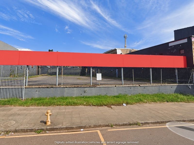 Street-level view of a commercial property featuring a paved lot, chain-link perimeter fencing, and a prominent red signage board on the building facade.