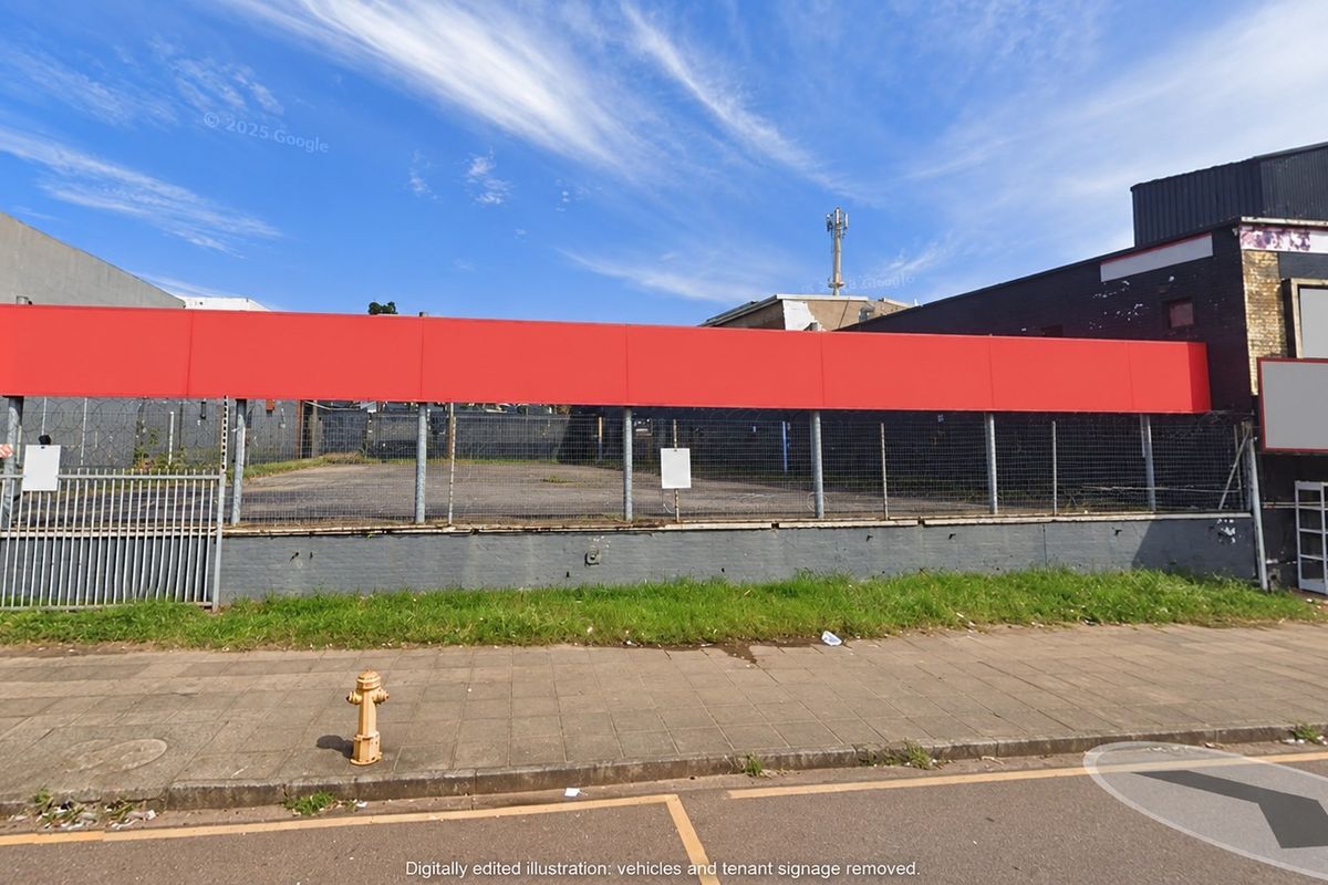 Street-level view of a commercial property featuring a paved lot, chain-link perimeter fencing, and a prominent red signage board on the building facade.
