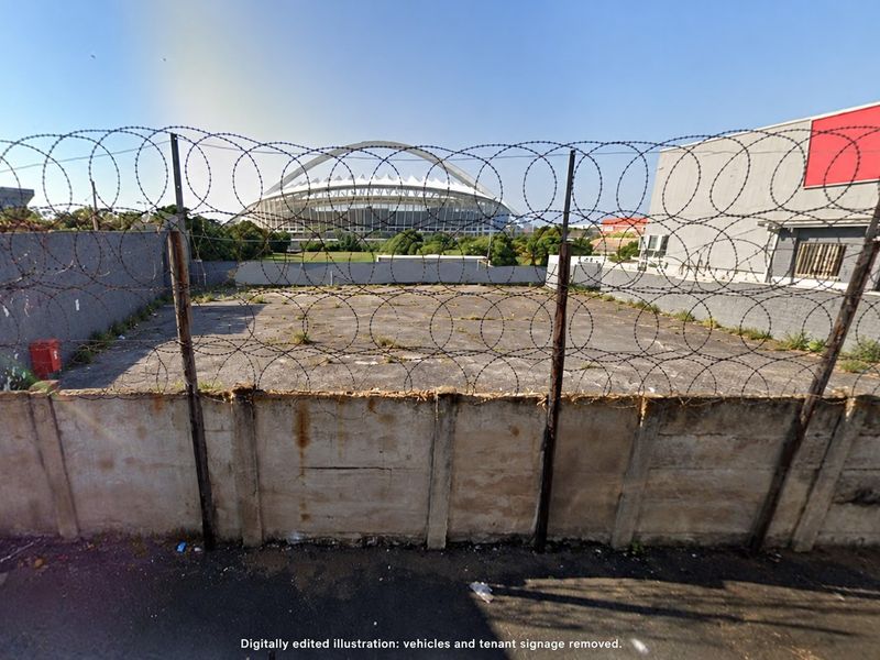 A vacant paved yard area enclosed by a concrete boundary wall and razor wire fencing, offering a view of the nearby stadium.