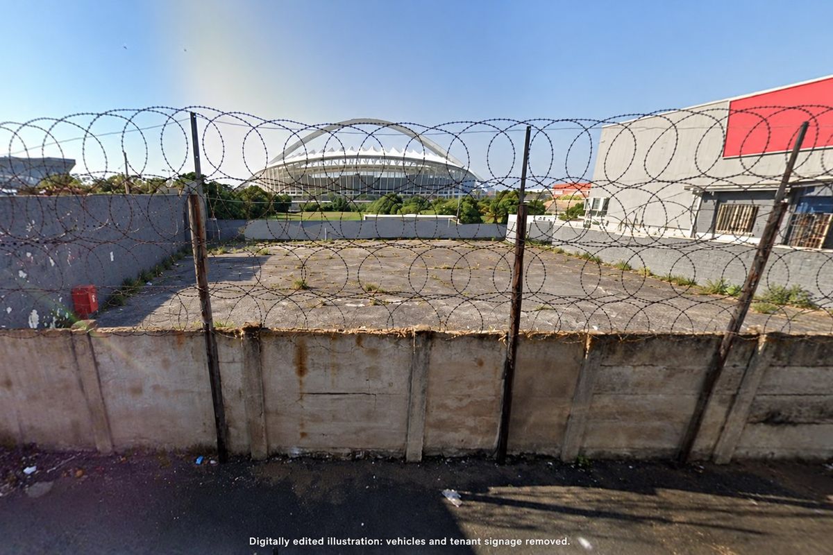 A vacant paved yard area enclosed by a concrete boundary wall and razor wire fencing, offering a view of the nearby stadium.