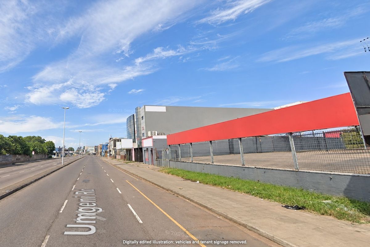 Street view of the commercial property frontage featuring a perimeter fence, retaining wall, and paved sidewalk along the main road.