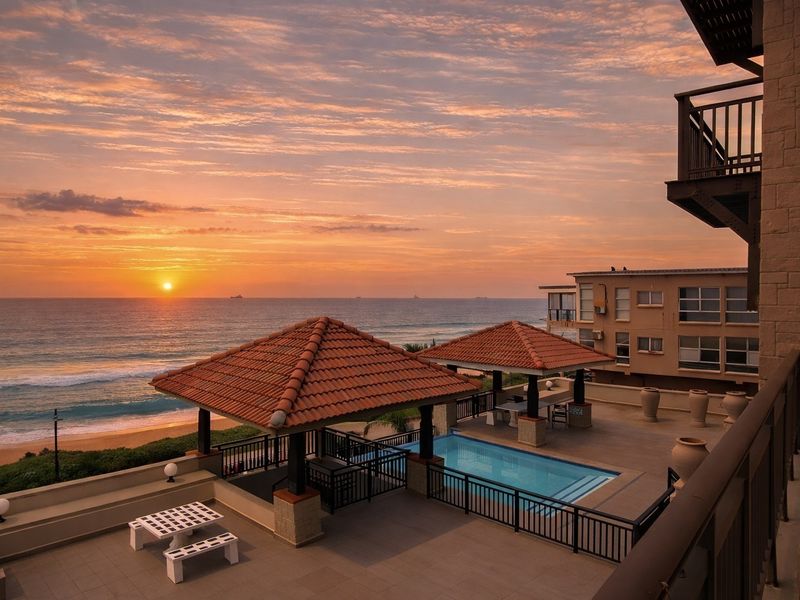 A stunning oceanfront pool area featuring tiled decking, covered gazebos, and direct views of the beach at sunset.